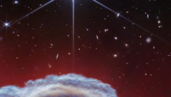 A clumpy dome of blueish-gray clouds rises about a third of the way from the bottom. Above it, streaky, translucent red wisps brush upward to about halfway up the image. The top half of the image is the black background of space with one prominent, bright white star with Webb’s 8-point diffraction spikes. Additional stars and galaxies are scattered throughout the image, although very few are seen through the thick clouds at bottom and all are significantly smaller than the largest star.