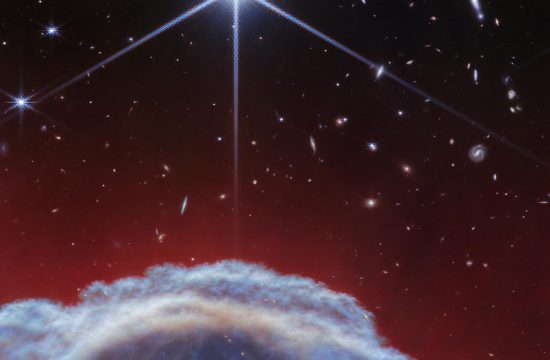 A clumpy dome of blueish-gray clouds rises about a third of the way from the bottom. Above it, streaky, translucent red wisps brush upward to about halfway up the image. The top half of the image is the black background of space with one prominent, bright white star with Webb’s 8-point diffraction spikes. Additional stars and galaxies are scattered throughout the image, although very few are seen through the thick clouds at bottom and all are significantly smaller than the largest star.
