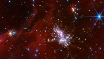 At center right is a compact star cluster composed of luminous red, blue, and white points of light. Faint jets with clumpy, diffuse material extend in various directions from the bright cluster. Above and to the right is a smaller cluster of stars. Translucent red wisps of material stretch across the scene, though there are patches and a noticeable gap in the top left corner that reveal the black background of space. Background galaxies are scattered across this swath of space, appearing as small blue-white and orange-white dots or fuzzy, thin disks. There are two noticeably larger points, foreground stars, with diffraction spikes: an orange-white point on the left, and a blue-white point in the top right.